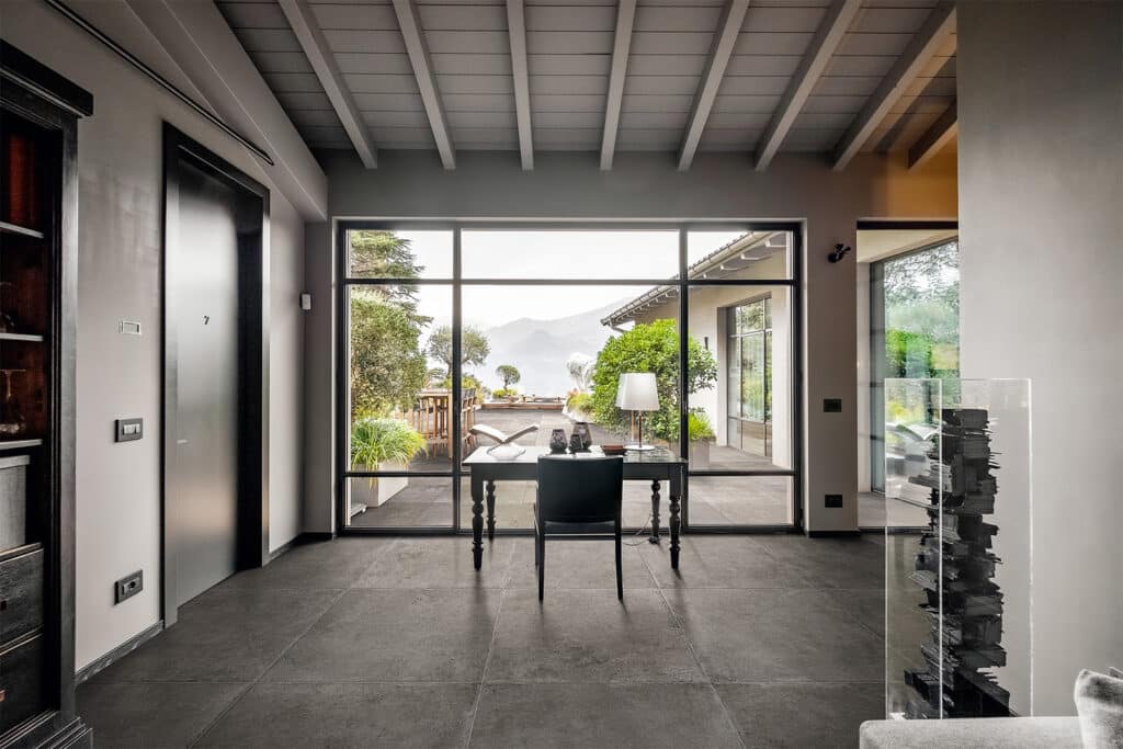 Intérieur moderne gris avec sol en carreaux foncés et plafond à poutres. Bureau simple devant une grande baie vitrée donnant sur un patio et des montagnes au loin. Porte marquée '7'.
