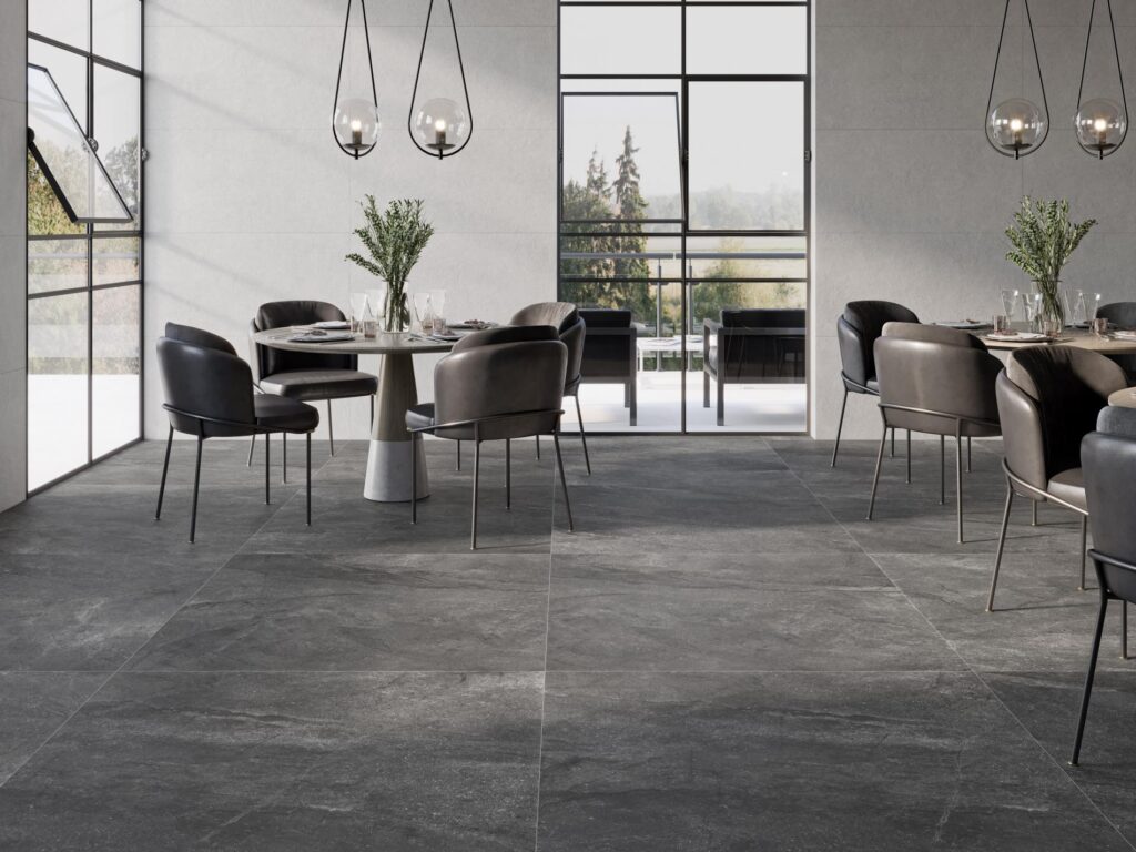 Salle à manger moderne avec sol en grands carreaux ardoise foncée, murs clairs, chaises en cuir noir et suspensions en verre au-dessus de tables rondes.