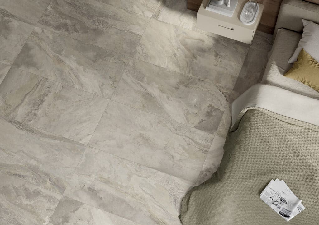 Vue en plongée sur un sol en grands carreaux de grès cérame effet marbre gris-beige à côté d'un lit moderne, près d'une table de chevet.