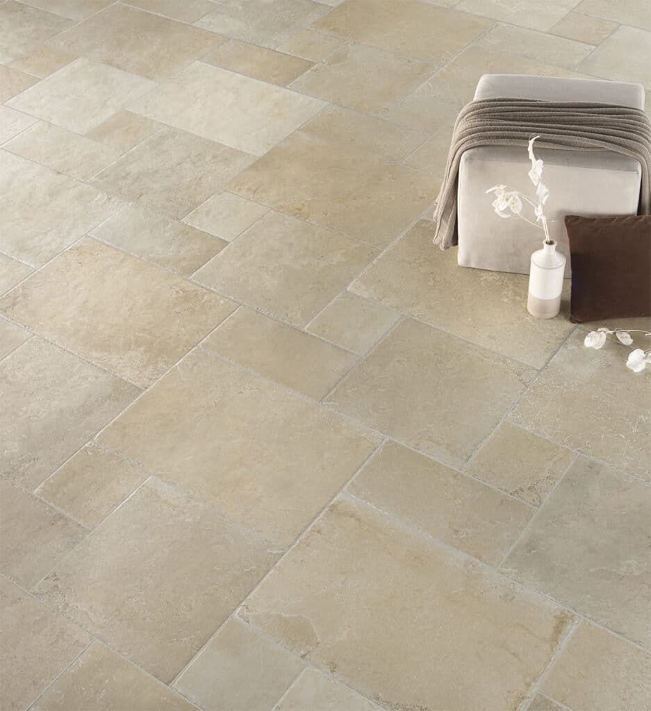 Sol en carreaux de pierre naturelle beige clair, style rustique, avec un pouf garni d'un jeté et un vase minimaliste.