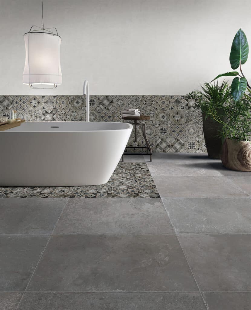 Salle de bain moderne avec baignoire îlot blanche, mur carrelé à motifs méditerranéens et grand pot de plantes vertes.