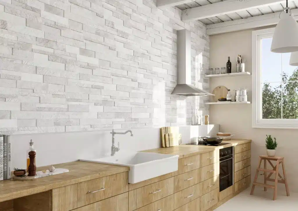 Cuisine moderne avec mur en pierre blanche texturée, comptoirs en bois clair, évier de ferme, hotte inox et étagères flottantes près d'une grande fenêtre.