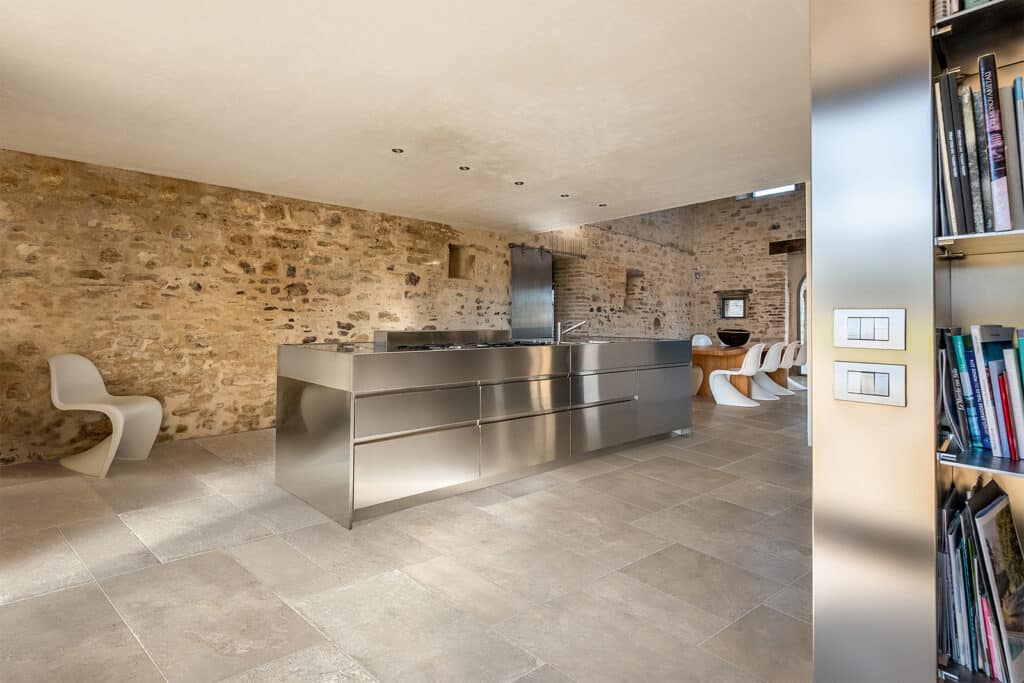 Cuisine moderne inox dans ancienne bâtisse pierre, sol carrelé beige, chaises design blanches, interrupteurs BTicino visibles.