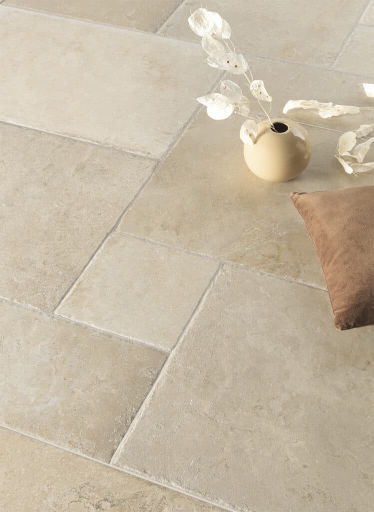 Sol en grandes dalles de pierre naturelle beige clair avec des joints gris, décoré d'un vase rond et d'un coussin marron.