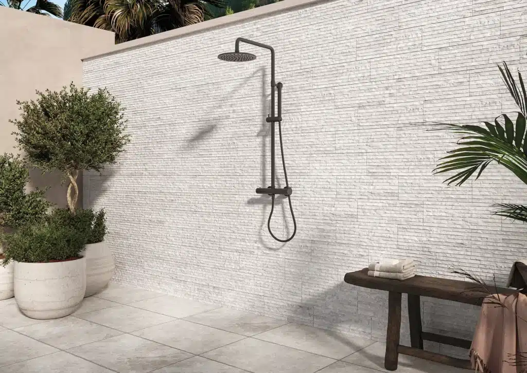 Douche extérieure moderne noire sur mur blanc texturé, avec plantes en potées et banc rustique, créant une ambiance spa.