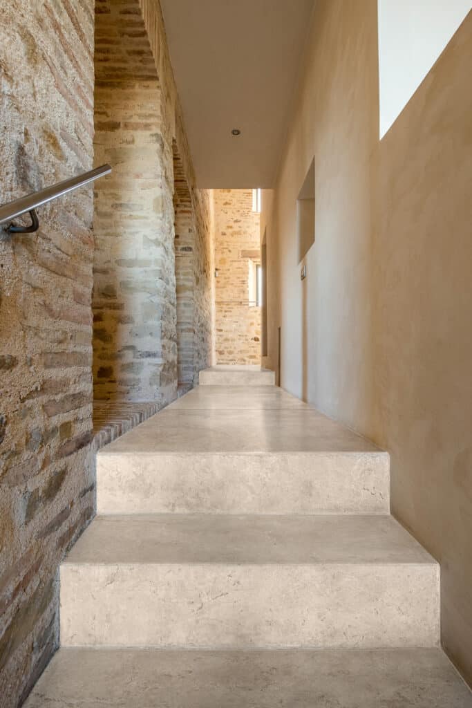 Escalier moderne en pierre beige montant dans un couloir étroit, contrastant avec un mur ancien en briques exposées et des murs lisses.