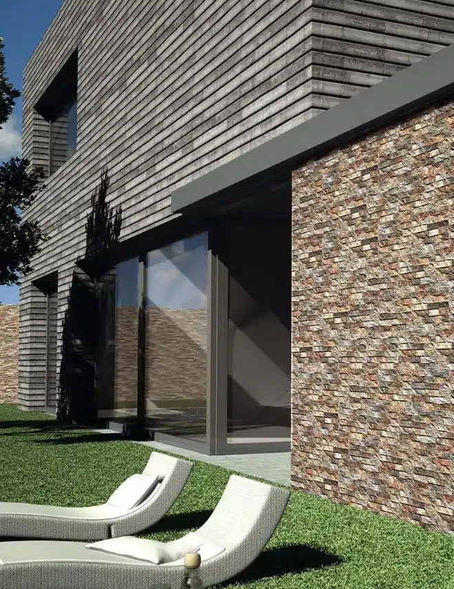 Façade moderne avec bardage gris horizontal contrastant avec un mur d'accent en pierre sèche multicolore, près de chaises longues sur pelouse verte.