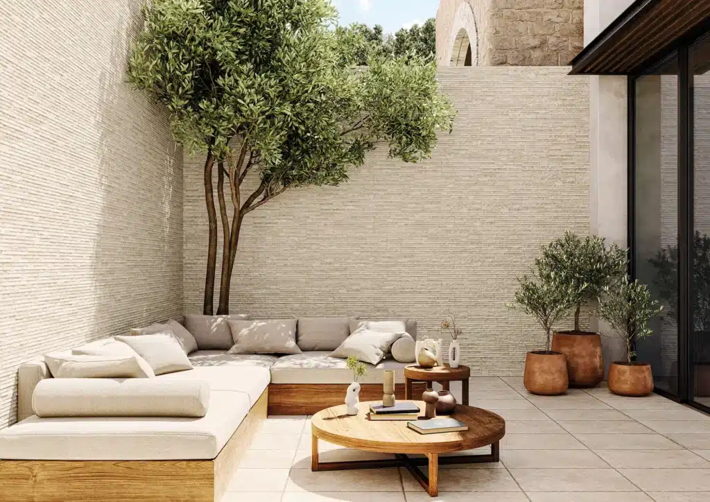 Patio extérieur moderne avec canapé d'angle en bois, coussins beiges, table basse ronde et arbre d'olivier sous murs texturés clairs.