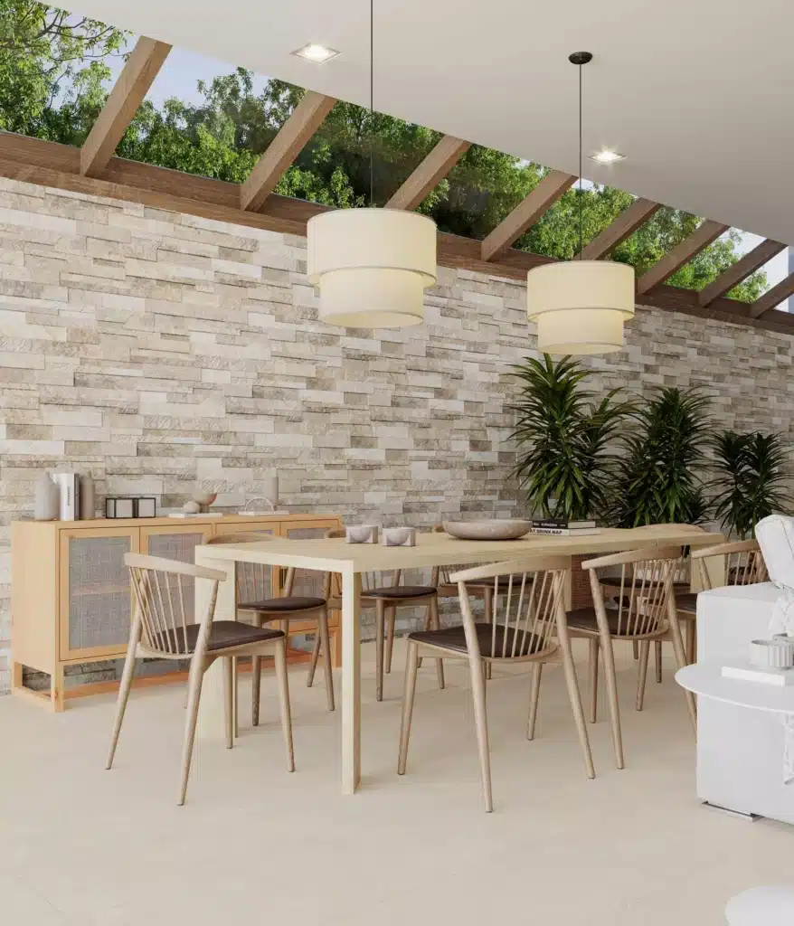 Salle à manger lumineuse avec mur de pierre claire, mobilier en bois clair, chaises à barreaux et suspension moderne sous une verrière laissant voir la verdure.