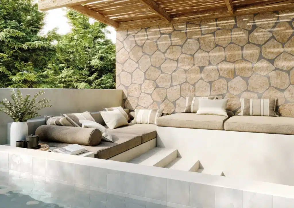 Coin salon extérieur luxueux près d'une piscine, avec des coussins beiges, un mur en pierres naturelles et une pergola en bois.