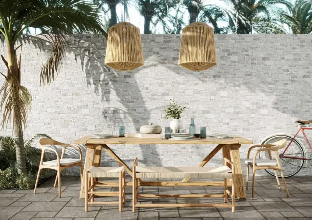 Salle à manger d'extérieur en bois clair avec suspensions corde, éclairée par le soleil, devant un mur de pierres blanches.