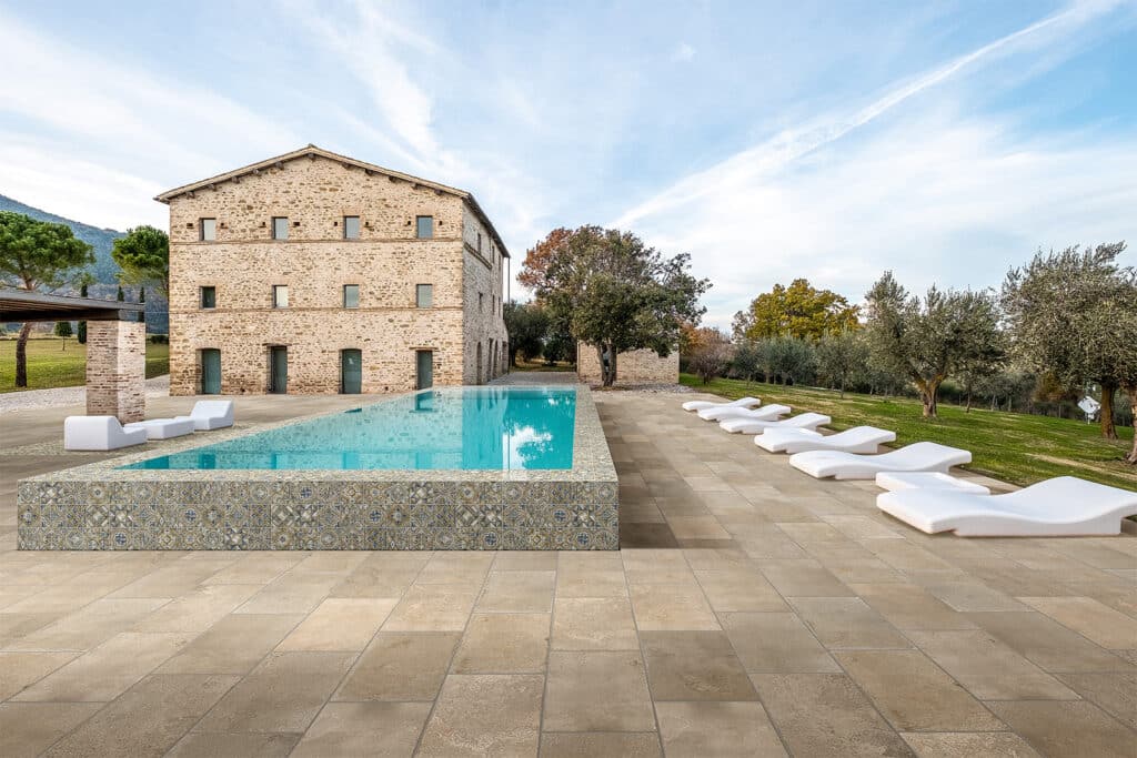 Piscine à débordement moderne et terrasse en dalles beiges devant une ancienne bâtisse en pierre toscane, avec chaises longues blanches.