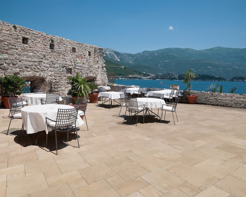 Terrasse de restaurant en pierre avec tables blanches et chaises en métal, vue sur une mer bleue et des montagnes verdoyantes sous un ciel clair.