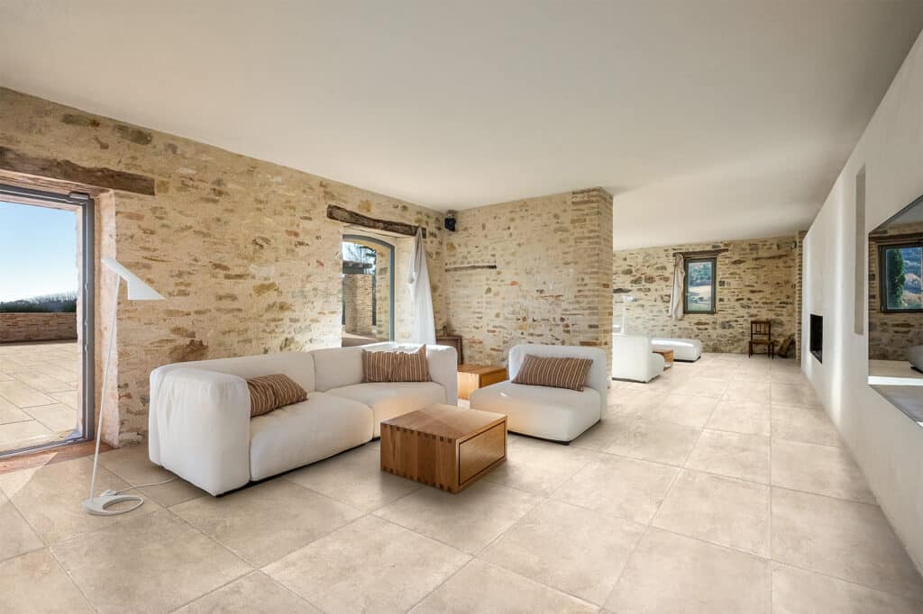 Carrelage salon rustique: murs en pierre et sol beige Salon lumineux aux murs en pierre et poutres apparentes. Sol carrelage intérieur extérieur beige, canapés blancs modernes.