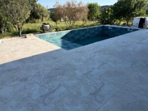 Piscine en cours de finition avec terrasse en pierre beige et mosaïque bleu canard, jardin en arrière-plan.