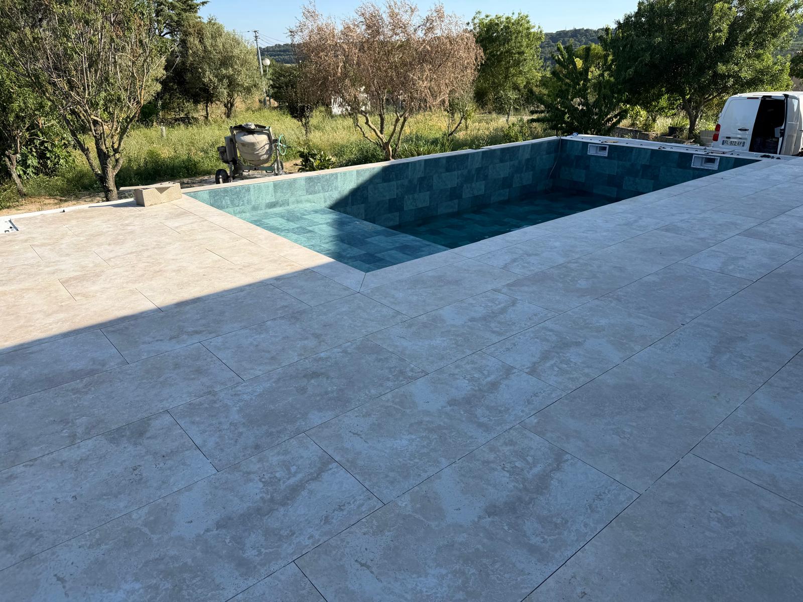 Piscine en cours de finition avec terrasse en pierre beige et mosaïque bleu canard, jardin en arrière-plan.