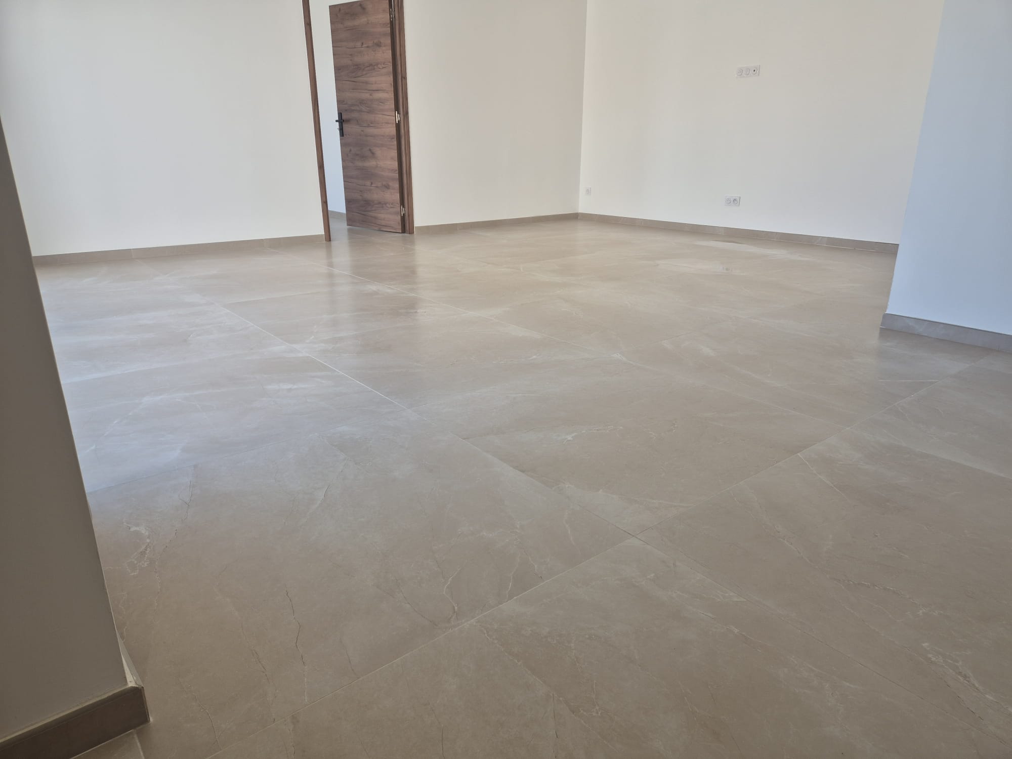 Carrelage grand format beige dans une pièce vide lumineuse avec porte en bois.