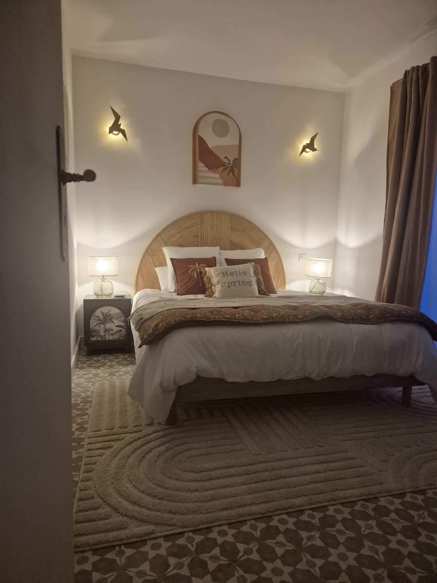 Chambre bohème avec tête de lit en rotin, lampes oiseaux et coussin 'Hello spring' sur tapis beige.