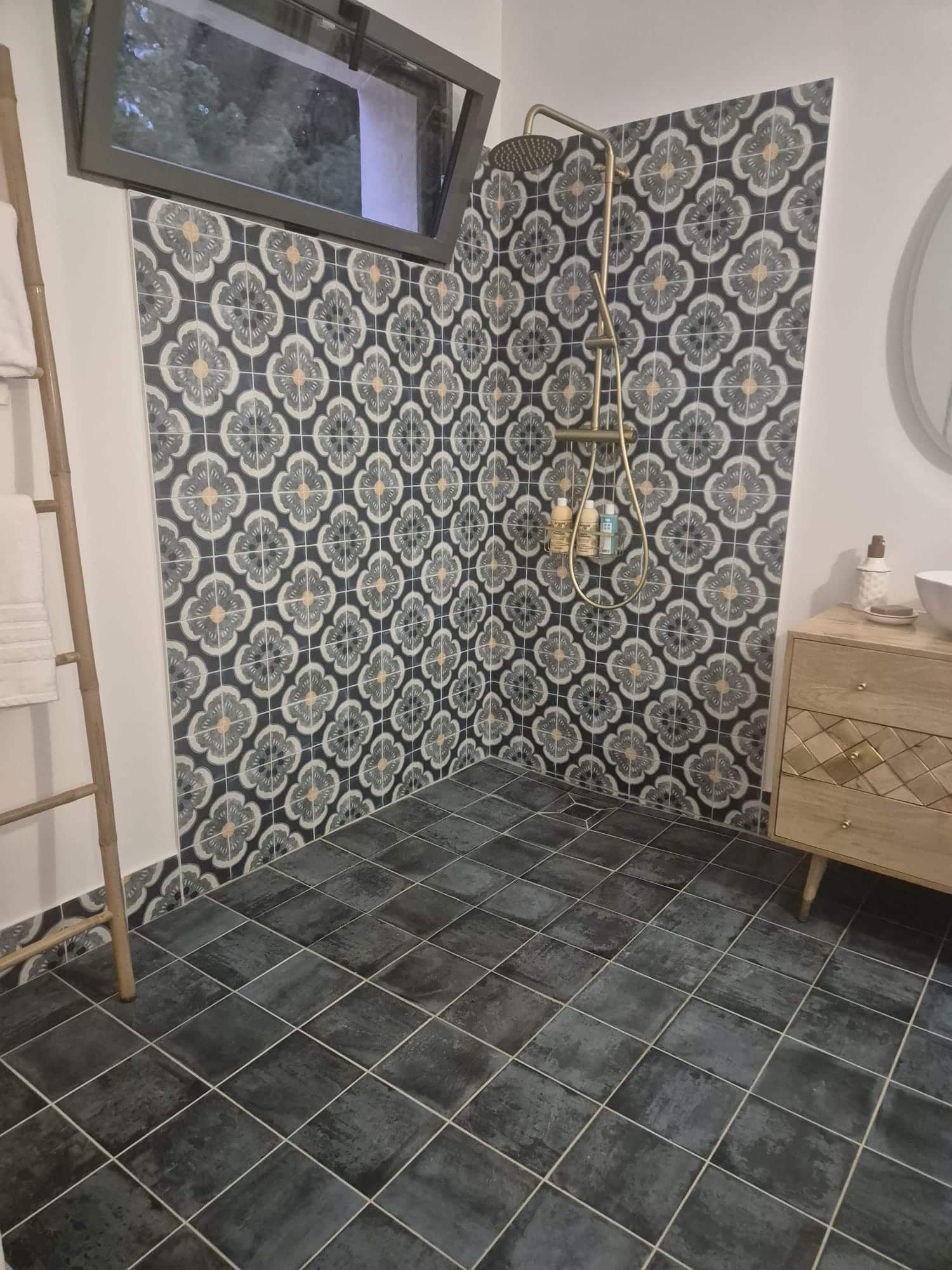 Douche italienne, carrelage à motifs gris et blanc, robinetterie dorée, meuble bois et échelle porte-serviette.