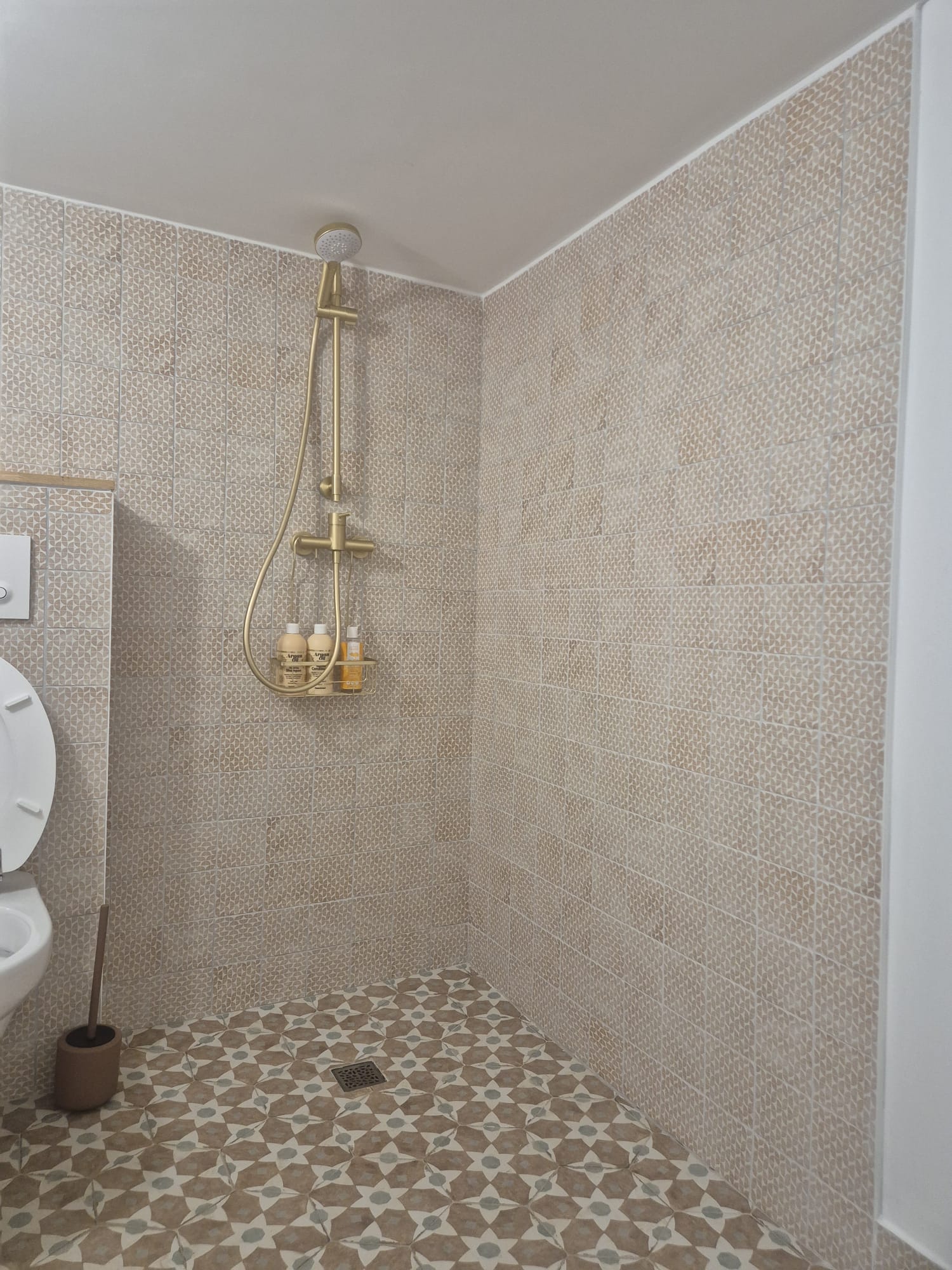 Douche avec colonne dorée, carrelage beige texturé et sol étoilé. Flacons de shampoing Argon Oil.