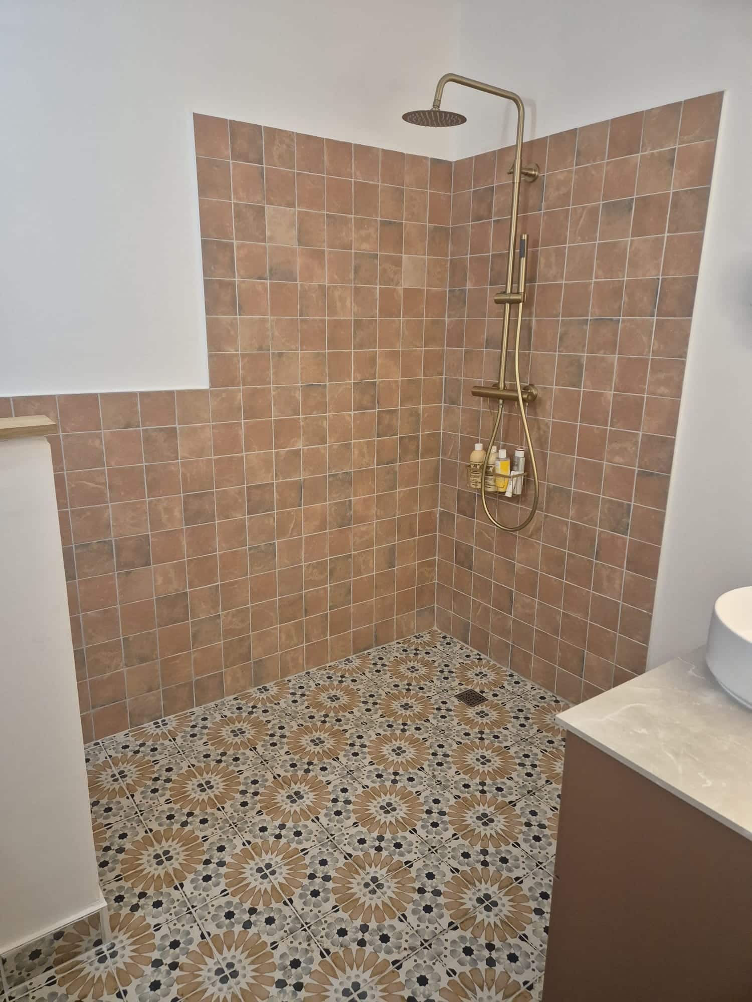 Douche italienne, carrelage terracotta, sol à motifs et robinetterie dorée.