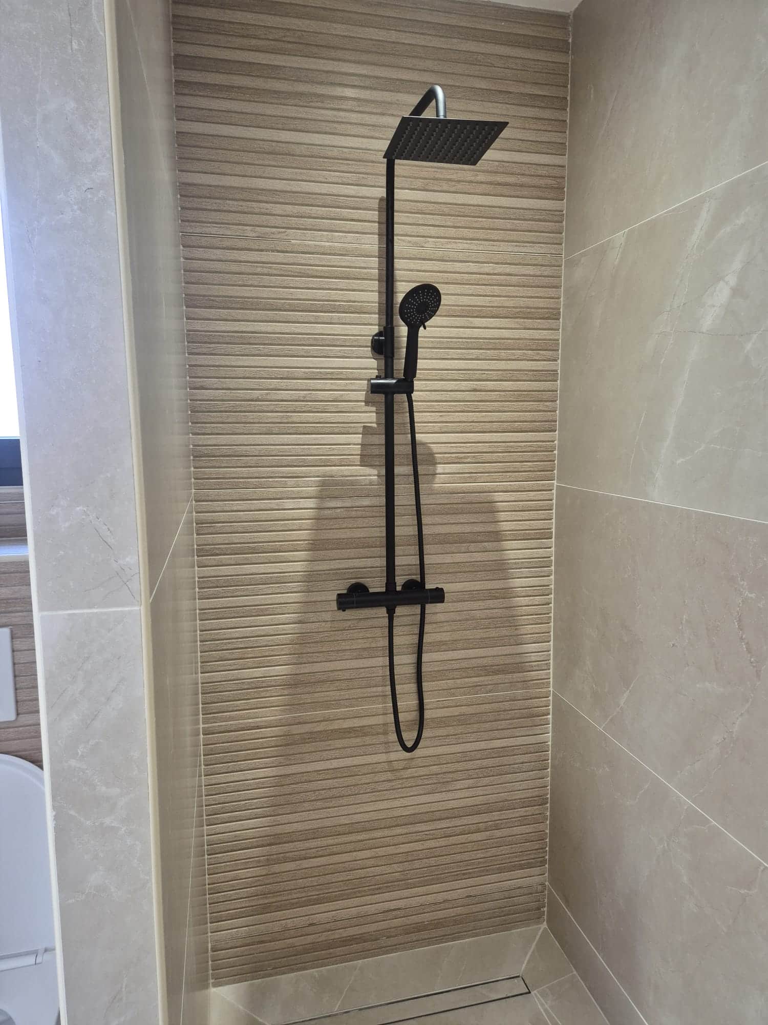 Douche noire mate sur carrelage beige strié et marbre.