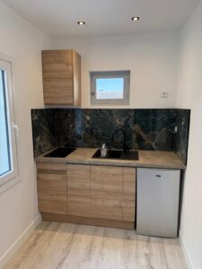 Kitchenette moderne avec meubles bois clair, crédence marbre sombre, évier noir et mini-réfrigérateur gris.