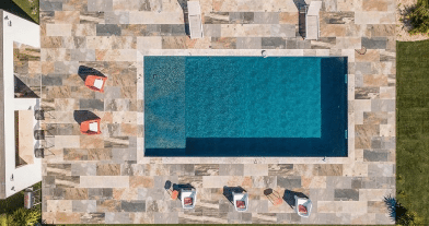 Vue aérienne d'une piscine bleue et terrasse en pierre naturelle avec chaises longues colorées.
