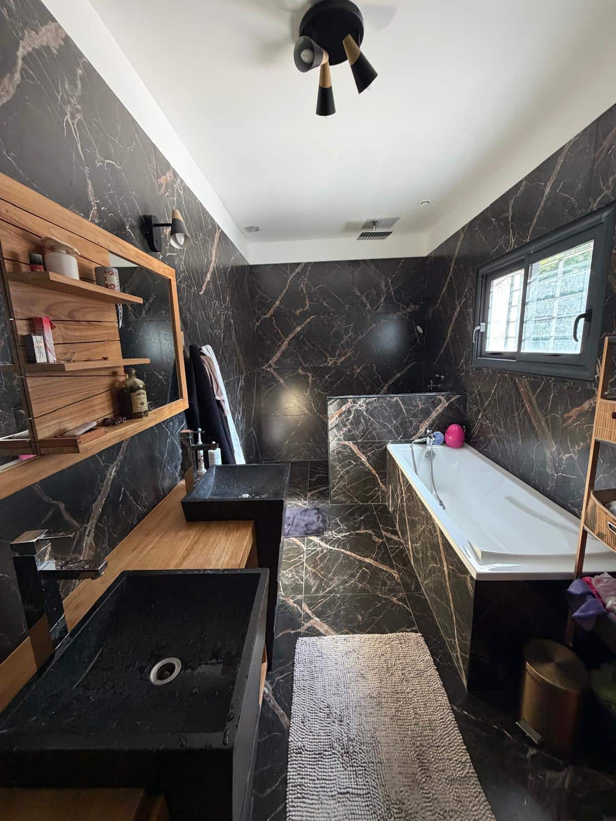 Salle de bain luxueuse en marbre noir avec double vasque noire, étagères en bois et baignoire blanche.