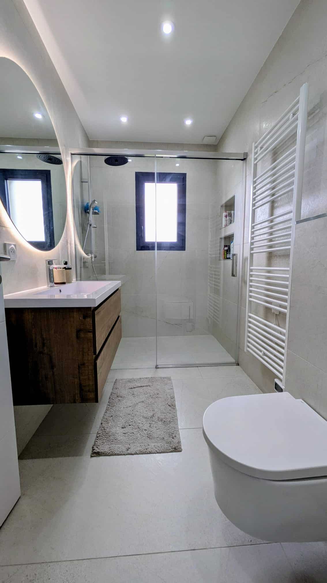 Salle de bain moderne avec meuble bois, miroir rétroéclairé, douche à l'italienne et WC suspendu blanc.