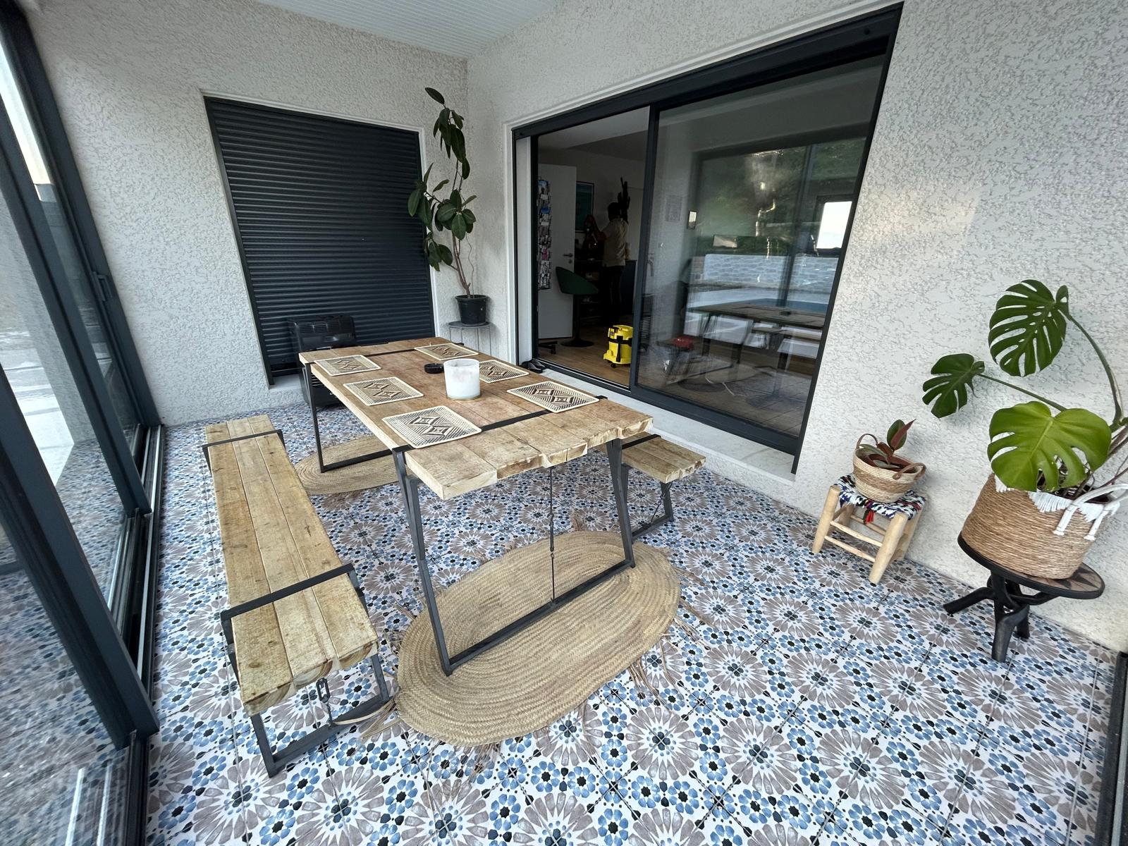 Terrasse avec table bois rustique, carrelage bleu à motifs et plantes Monstera sous verrière moderne.